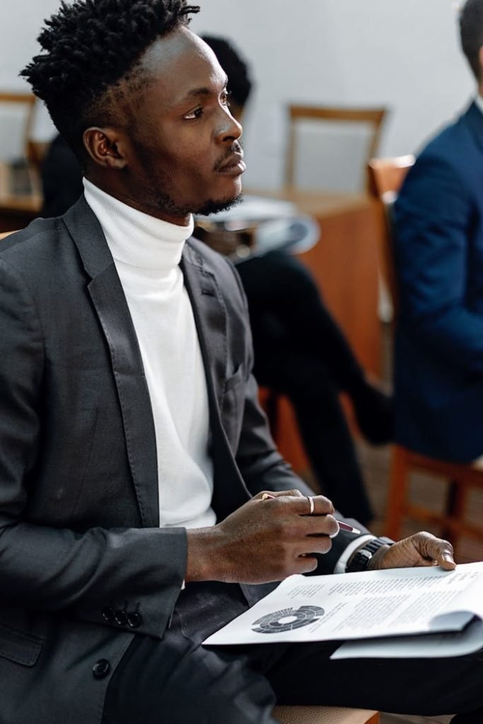 Young professional in black suit attentively listening during a presentation, holding documents.