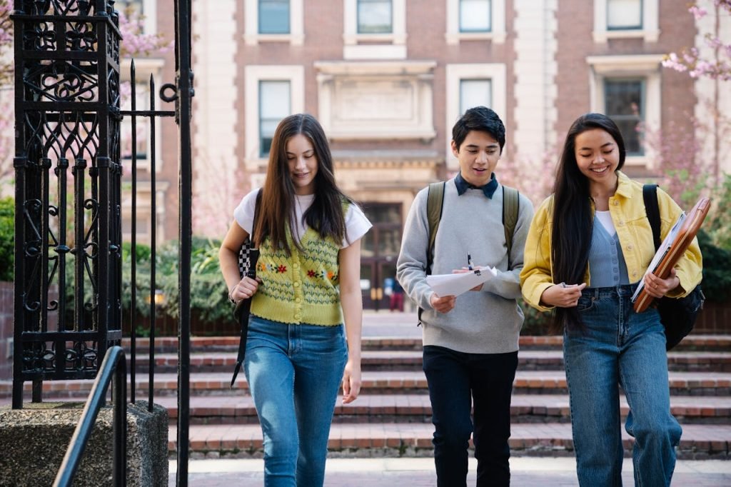 Three smiling students walking together on a university campus, holding notebooks and backpacks.